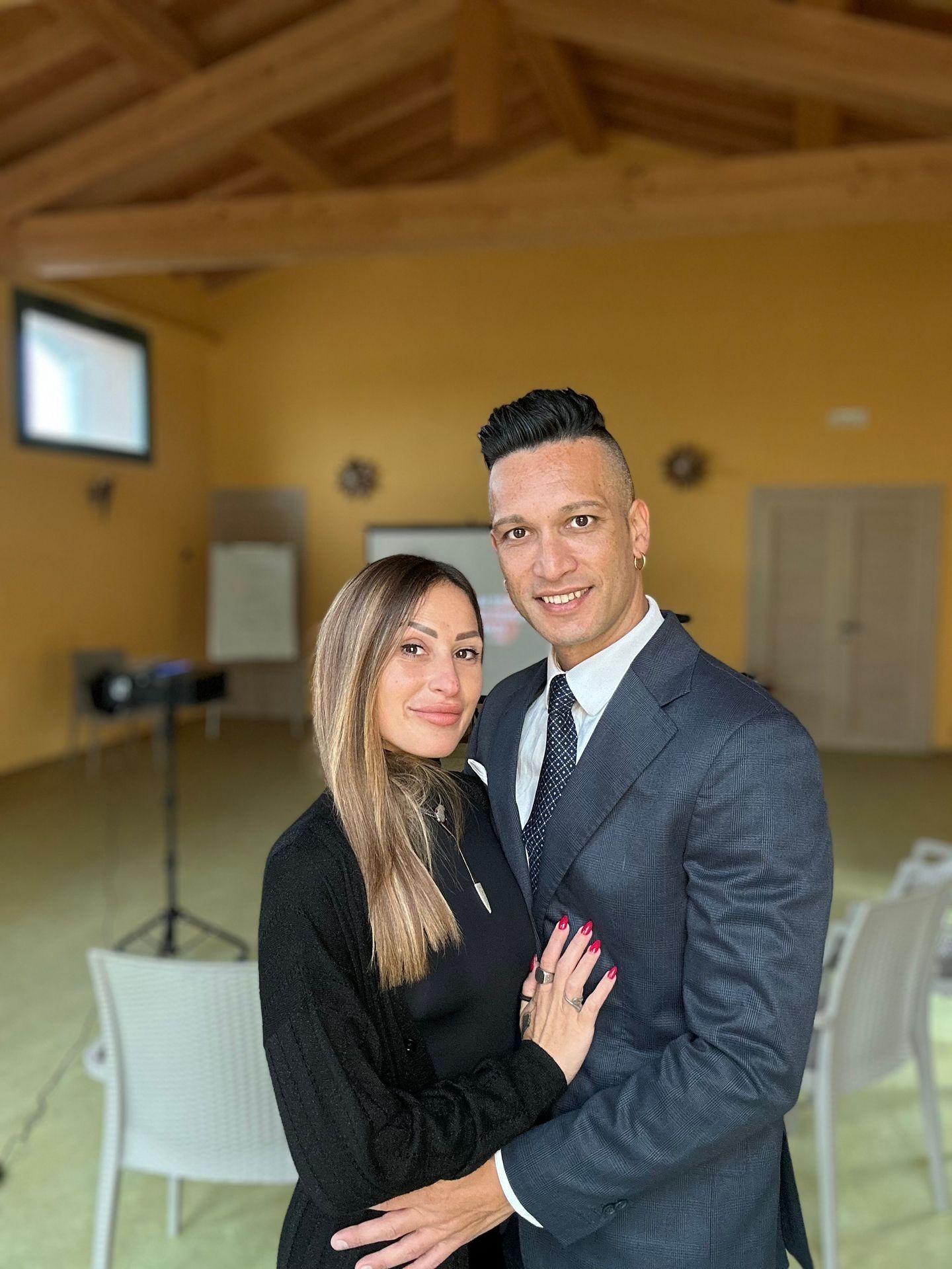 Couple posing together in an indoor setting with wooden ceiling and chairs in the background.