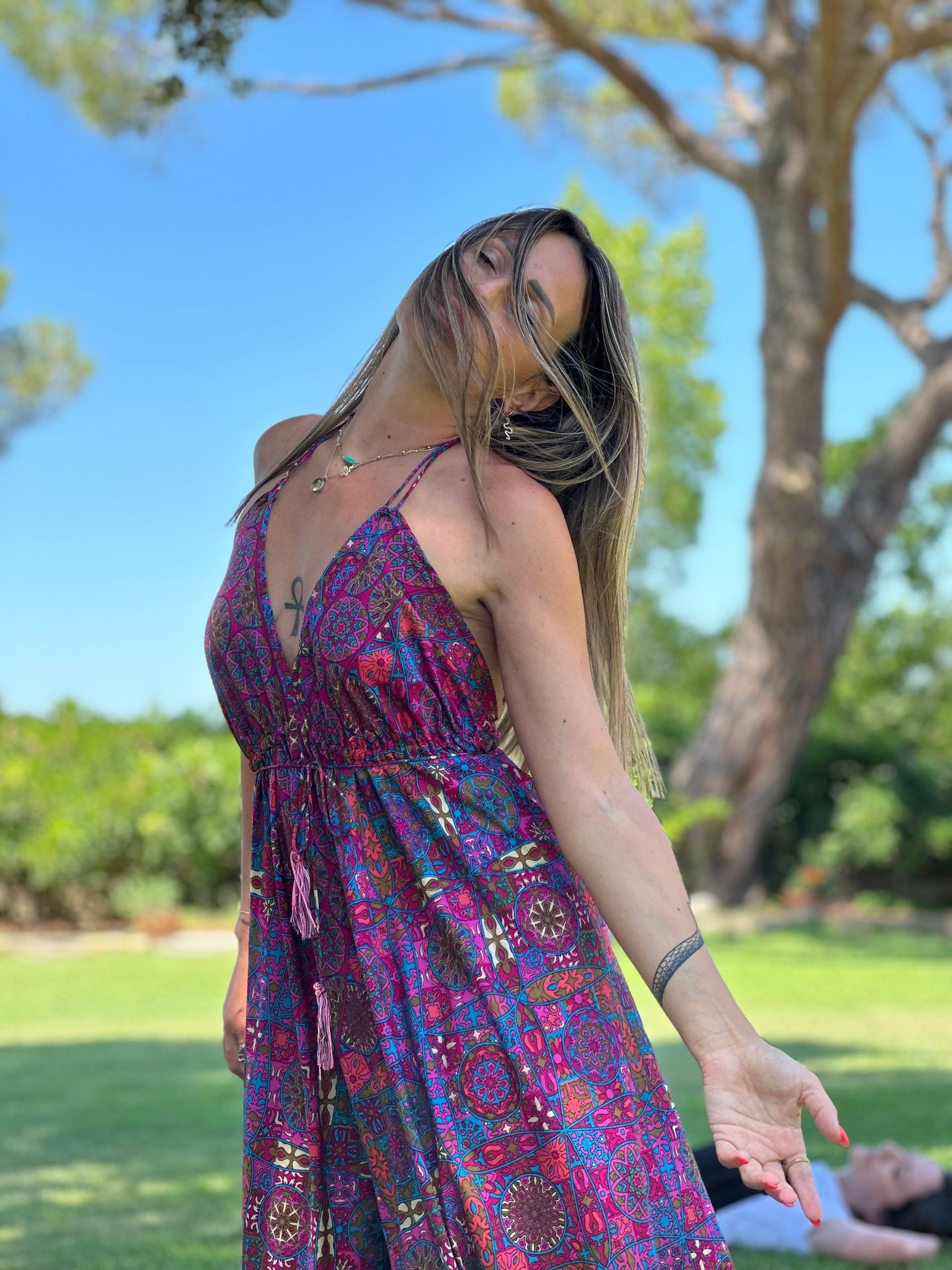 Woman in a colorful patterned dress posing outdoors with trees and grass in the background.