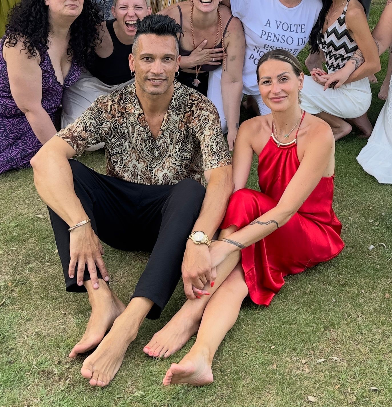 Group of people sitting on grass, two in foreground smiling, others posing and laughing behind.